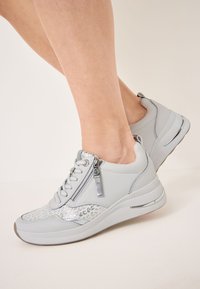 Sneaker grigie chiare con un accento argento testurizzato, dotate di dettagli con zip e lacci, su una suola bianca spessa con una striscia metallica.