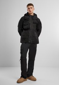 Giacca utility nera con più tasche, tessuto texturizzato, bottoni a pressione e cappuccio, abbinata a pantaloni cargo scuri e scarpe chunky color sabbia.