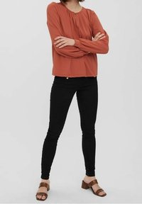 Blouse couleur rouille avec un col rond et des détails froncés, associée à un jean skinny noir et des sandales à lanières marron.