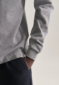 Grau Sweatshirt mit strukturiertem Finish und gerippten Bündchen, getragen über dunklen Hosen mit einer Hand, die in der Tasche ruht. Schlichtes, modernes Design.
