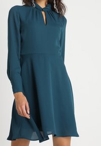 Teal mekko kevyestä kankaasta, jossa on avainaukko-olkaimet, pitkät hihat ja liehuva hame, jossa on A-linjainen siluetti.