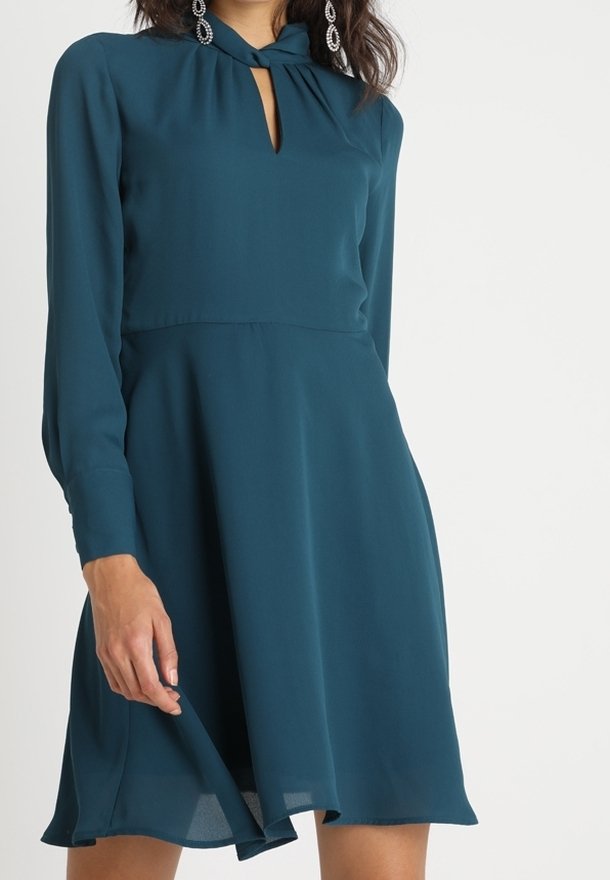 Teal mekko kevyestä kankaasta, jossa on avainaukko-olkaimet, pitkät hihat ja liehuva hame, jossa on A-linjainen siluetti.