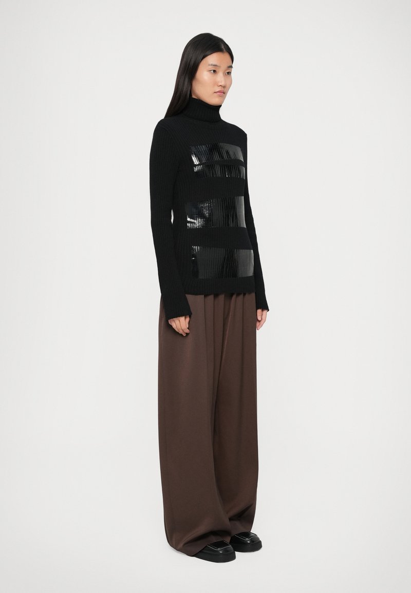 Pull en laine côtelée noir à col roulé avec des panneaux horizontaux brillants noirs, associé à un pantalon brun ample et des chaussures plates noires.