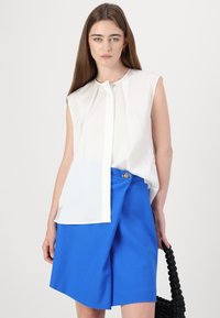 Witte, mouwloze top met een geplooide voorkant, gecombineerd met hooggevoerde, felblauwe shorts met een decoratieve knop en een gestructureerde stof.