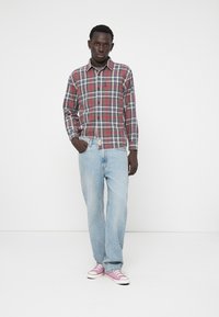 Chemise à carreaux rouge et bleu avec des manches longues, associée à un jean bleu clair à jambes larges et des baskets roses. Le modèle se tient avec une main dans la poche.