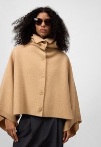 Poncho beige in lana con colletto alto, caratterizzato da grandi bottoni sul davanti. Texture morbida e maniche ampie, abbinato a pantaloni grigio scuro.