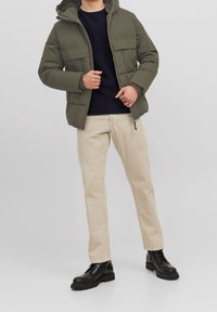 Giacca imbottita verde oliva con cappuccio, due tasche frontali, indossata sopra un maglione lavorato a maglia blu navy, abbinata a pantaloni beige e stivali neri con lacci.