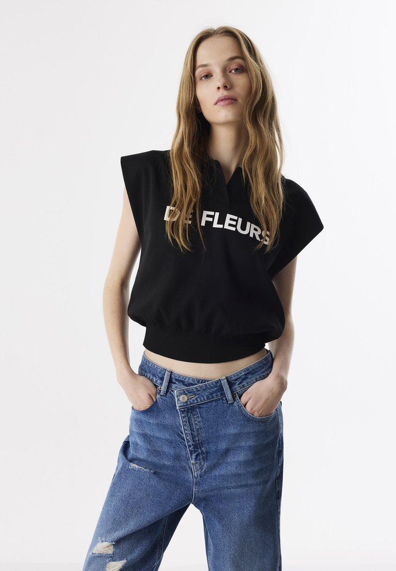 Felpa cropped nera con testo "DE FLEURS", maniche corte, abbinata a jeans in denim blu a vita alta con dettagli sfilacciati.