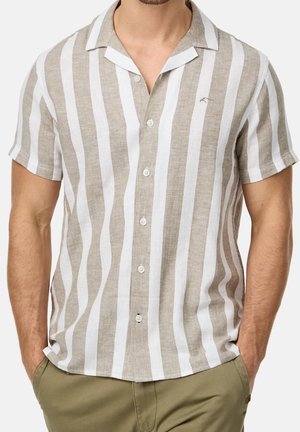 Mann trägt ein kurzärmliges, beige-weiß vertikal gestreiftes Hemd mit Knopfleiste und Kragen, Hände in den Taschen olivgrüner Hosen.