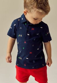 Marineblå polo med flerfarvede broderede mønstre af biler, træer og dyr; sammen med lyse røde shorts. Korte ærmer og krave.