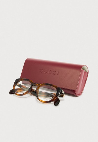 Ochelari Gucci cu un model de țestoasă, rame negre groase și lentile transparente, acompaniați de un capac dur maron închis, embosat cu "GUCCI."
