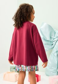 Bordeauxrotes übergroßes Sweatshirt mit langen Ärmeln, getragen über einem bunten, mit Pailletten besetzten Rock. Weiche Textur, legerer Sitz, präsentiert vor pastellfarbenem Hintergrund.