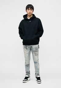 Must kapuutsiga dressipluus ees taskuga, kombineeritud helesiniste kulunud teksadega, millel on graafilised trükid, ning mustade ja valgete tossudega.