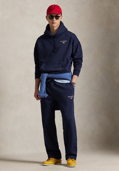 Sweat à capuche et pantalon de survêtement bleu marine avec logo « Polo Sport » ; pull bleu clair noué à la taille ; casquette rouge ; chaussures jaunes. Texture de tissu lisse.
