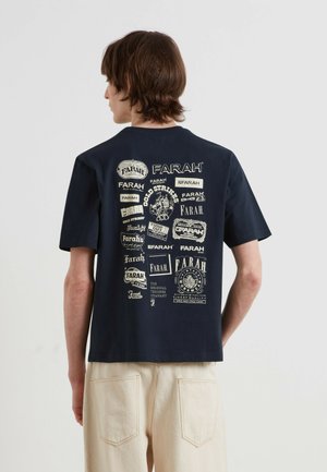 Junge Person mit kurzen braunen Haaren trägt ein marineblaues T-Shirt mit mehreren weißen Farah-Markenlogos auf dem Rücken und hellbeige Hosen.