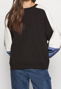 Sudadera negra con acentos en paneles blancos y azules en las mangas, presenta un cuello redondo y puños acanalados. Logotipo bordado en la parte frontal.