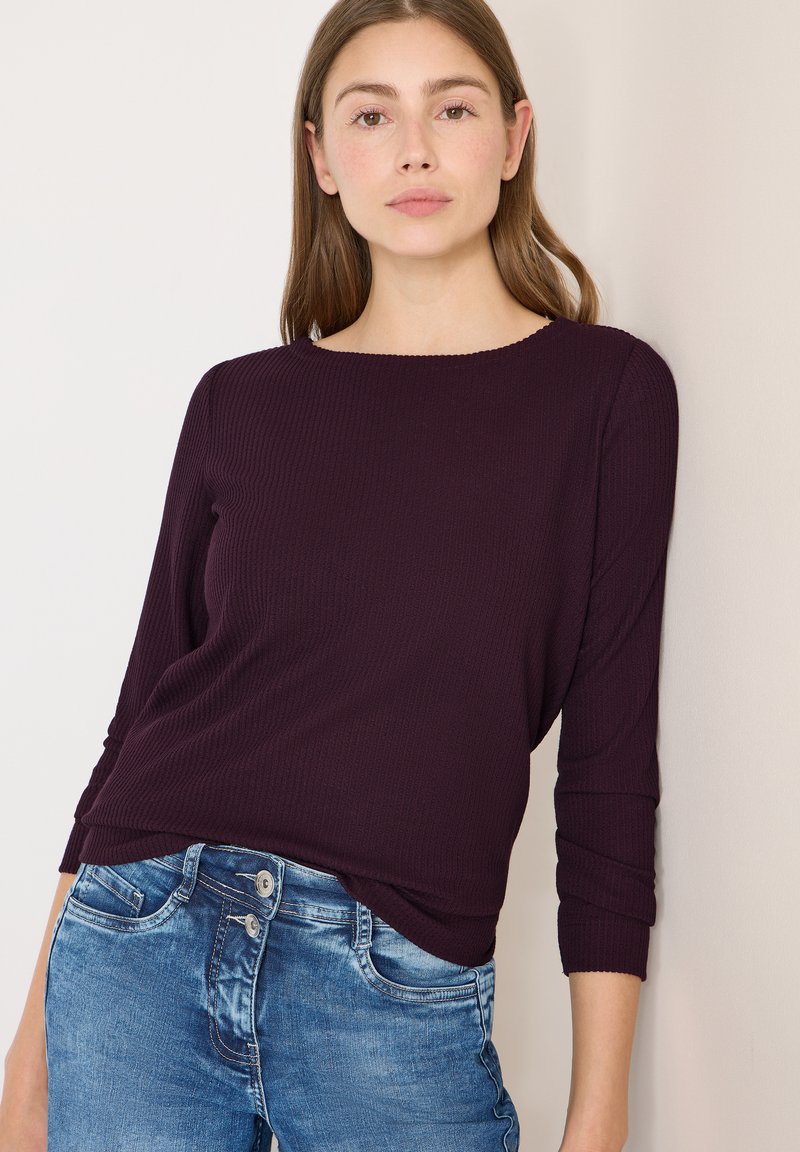 Tumma purppuranvärinen, ribbineulottu pitkähihainen paita venepääntiellä, yhdistetty vaaleansinisiin farkkuihin, joissa on kaksoisnapitus.