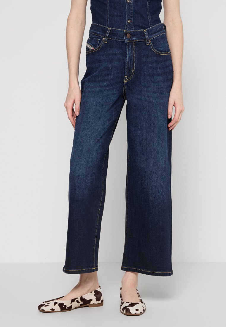 Dark blue wide-leg jeans with a high waist, featuring front pockets and a button closure; worn with cream flats featuring a cow print.