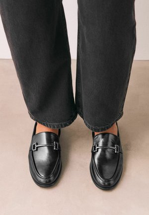 Mocasines negros de cuero con detalle de pieza metálica, usados con pantalones negros de pierna recta sobre un suelo beige.