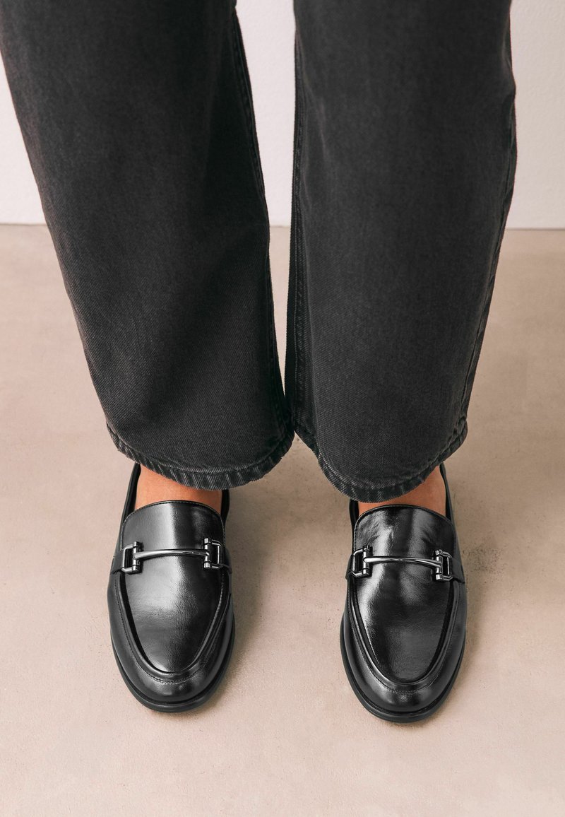 Mocassins en cuir noir avec détail en métal, portés avec un pantalon noir à jambes droites sur un sol beige.