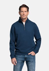 Donkerblauwe zip-up trui met hoge kraag, textuurstof en lange mouwen, gecombineerd met lichtblauwe jeans. Heeft een ritssluiting aan de voorkant.