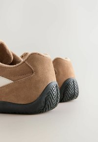 Chaussures en daim marron avec semelles en caoutchouc noir. Forme arrondie, texture douce et accents blancs contrastants sur les côtés. La vue arrière montre le motif de la semelle.
