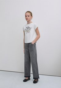 Maglietta grafica bianca a maniche corte con design colorato di un'aquila. Jeans grigi a gamba larga con decorazioni sparse e ballerine nere.