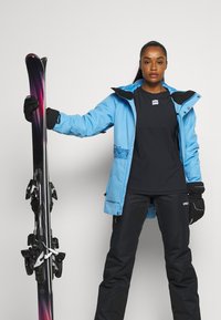 Blå skijakke med svarte aksenter, svart skjorte og svarte skibukser. Holder ski med lilla og svart gradientdesign, utstyrt med bindinger.