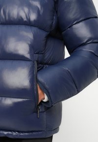 Námornícka puffer bunda s lesklým povrchom, obsahujúca zipsovú kapsu a elastické manžety. Prešívaný dizajn poskytuje izoláciu.
