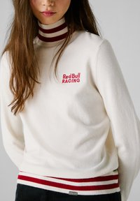 Maglione in maglia crema chiara con un alto colletto a righe rosse e bianche, con "Red Bull RACING" ricamato in rosso. Orlo a coste rosso e bianco.