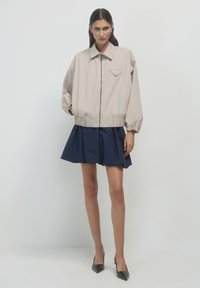 Beige Reißverschlussjacke mit spitzem Brusttasche, elastischen Bündchen, getragen über einem marineblauen ausgestellten Rock. Das Modell steht mit den Händen in den Taschen.