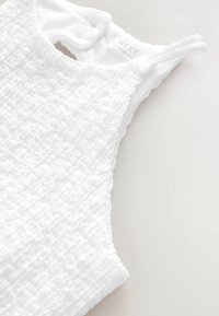 Vit ärmlös topp med en texturerad yta, med rund hals och öppning med nyckelhål på baksidan. Etikett synlig på innersömmen.