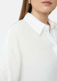 Biała koszula z fakturowanej tkaniny z klasycznym kołnierzykiem, z zapięciem na guziki z przodu i subtelnymi detalami na materiale. Okrągły dół i miękki, luźny krój.