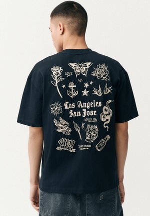 Crna pamučna majica s bijelim grafičkim tetovažama na leđima, uključujući cvijeće, sidro i tekst "Los Angeles San Jose." Kratki rukavi, ležeran kroj.