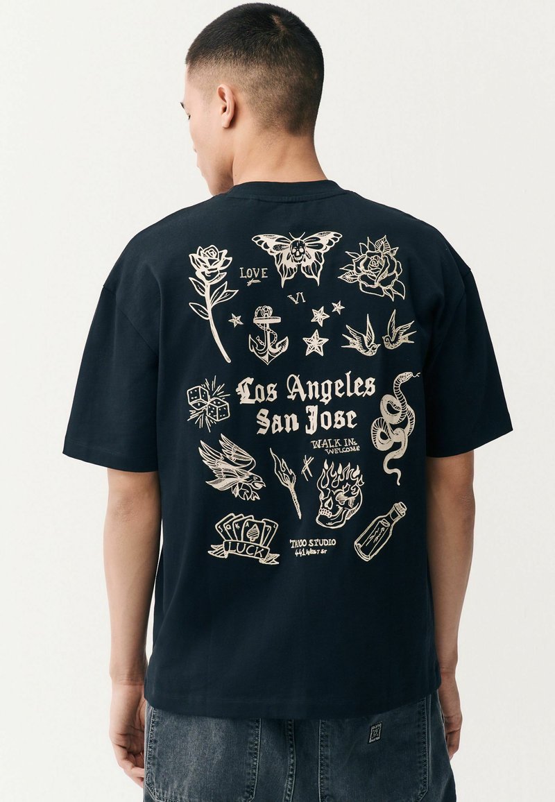 Crna pamučna majica s bijelim grafičkim tetovažama na leđima, uključujući cvijeće, sidro i tekst "Los Angeles San Jose." Kratki rukavi, ležeran kroj.