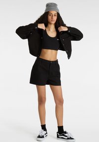 Giubbotto di denim nero sopra un top corto nero e pantaloni corti neri. Maniche corte, chiusura con bottoni a scatto e branding minimale. Sneakers nere con dettagli bianchi.