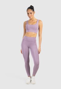 Reggiseno sportivo lilla e leggings a vita alta in un tessuto liscio e elastico. Il modello indossa scarpe da ginnastica bianche. Sfondo neutro.