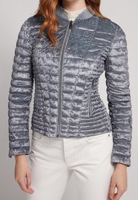 Femme portant une veste en velours matelassée argent-gris ajustée, avec une fermeture éclair à l'avant et des poches latérales zippées, associée à un pantalon blanc.