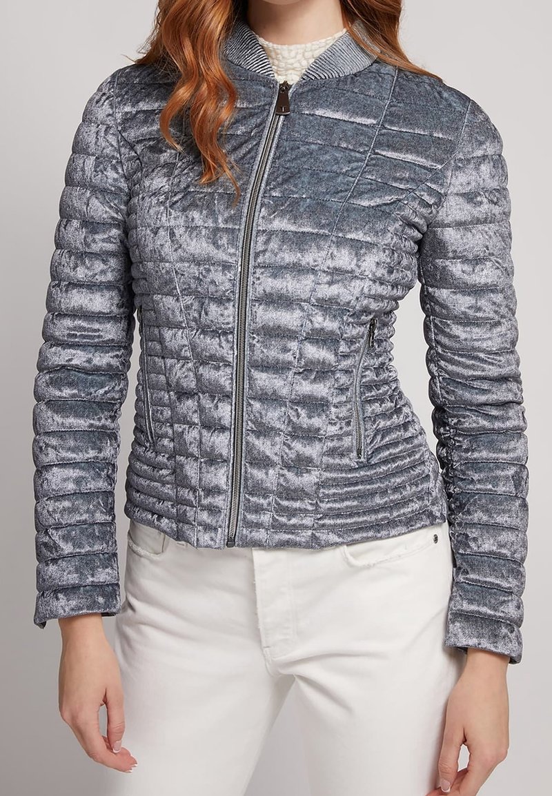 Femme portant une veste en velours matelassée argent-gris ajustée, avec une fermeture éclair à l'avant et des poches latérales zippées, associée à un pantalon blanc.