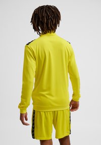 Lysegul atletisk top med lange ærmer, lavet i et struktureret stof med sorte detaljer på skuldrene. Kombineret med matchende shorts.