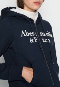 Felpa con zip di colore blu navy, realizzata in tessuto morbido, con tasca frontale e logo ricamato bianco, cappuccio foderato e polsini a costine.