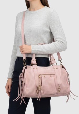 Donna che tiene una borsa in camoscio rosa chiaro con doppi manici, tracolla regolabile, cerniera e dettagli con nappine, indossa un maglione grigio e pantaloni neri.