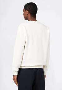 Maglione bianco con una texture liscia, colletto a girocollo e polsini a costine. Abbinato a pantaloni scuri, visto da dietro. Design minimalista.