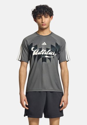 Giovane uomo con capelli ricci che indossa una maglietta grigia Adidas con stampa geometrica nera e pantaloncini neri, in piedi davanti a uno sfondo neutro.