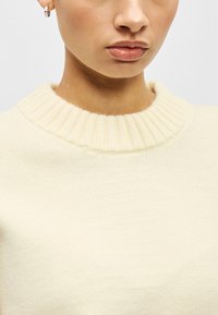 Πουλόβερ σε μπεζ απόχρωση με ριμπ ύφασμα και στρογγυλή λαιμόκοψη. Απαλό, υφασμένο ύφασμα με υφή. Κοντινή λήψη που αναδεικνύει το γιακά και την περιοχή του πάνω μέρους του στήθους.