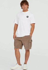 Tânăr stand cu mâinile în buzunare, purtând un tricou alb, pantaloni scurți maro și adidași albi, pe un fundal neutru.