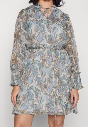 Woman wearing a knee-length, long-sleeve dress with a blue and brown paisley pattern, ruffled cuffs, and a tie neckline.