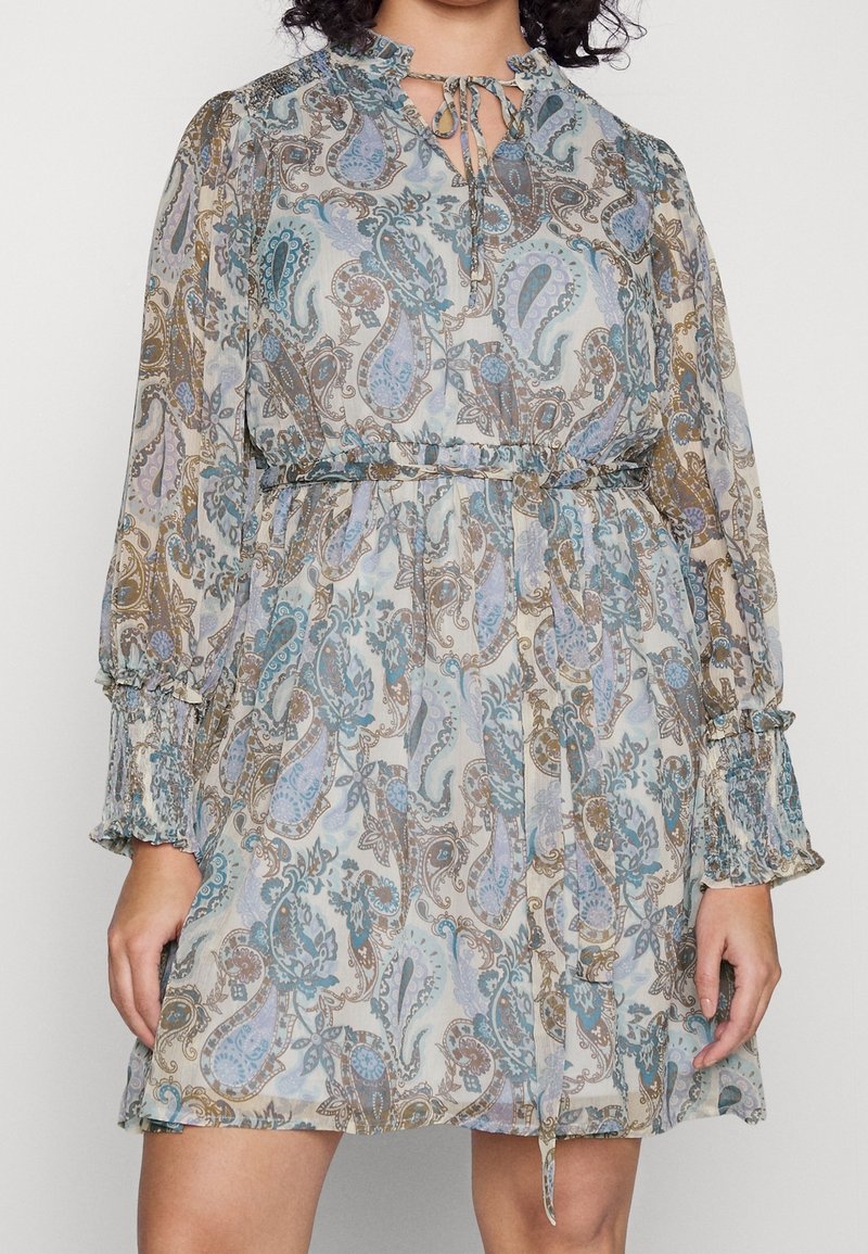 Femme portant une robe longueur genou, manches longues, avec un motif cachemire bleu et marron, des poignets volantés et un col à nouer.