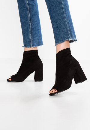 Person trägt gekürzte blaue Jeans und schwarze Wildleder-Stiefeletten mit offenen Zehen und hohen Absätzen auf weißem Boden.