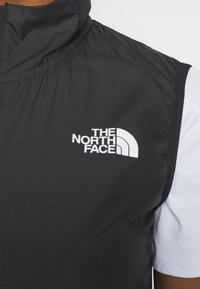 Svart ärmlös väst med hög krage, prydd med en vit The North Face-logotyp på framsidan, med en slät textur och en lätt design.
